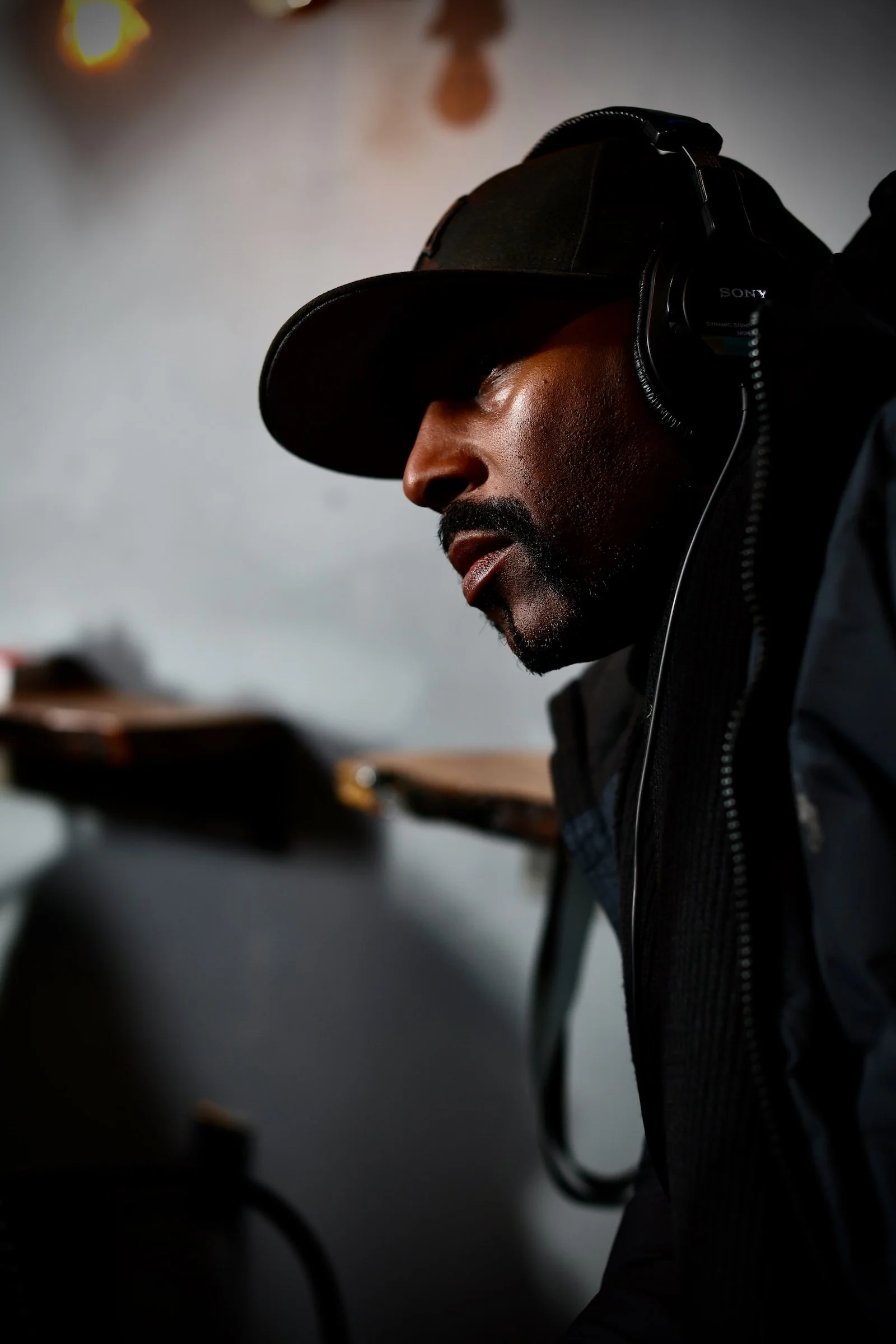 A man wearing a black cap, black jacket, and Sony headphones, sitting and looking downward, with a blurred background.