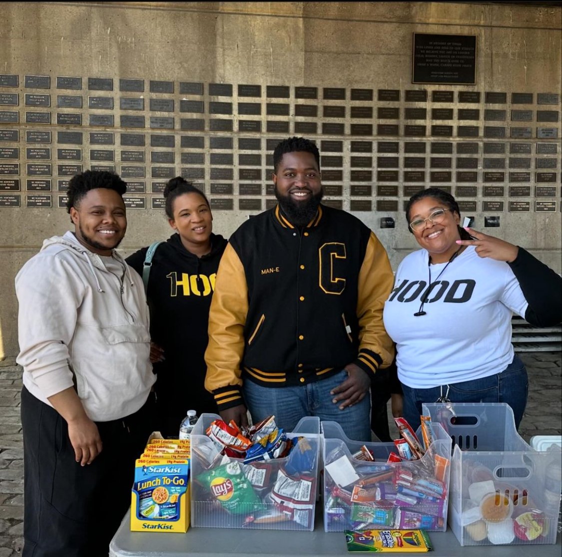 Four people smiling and posing in front of a display table with snack foods, standing against a wall with plaques.