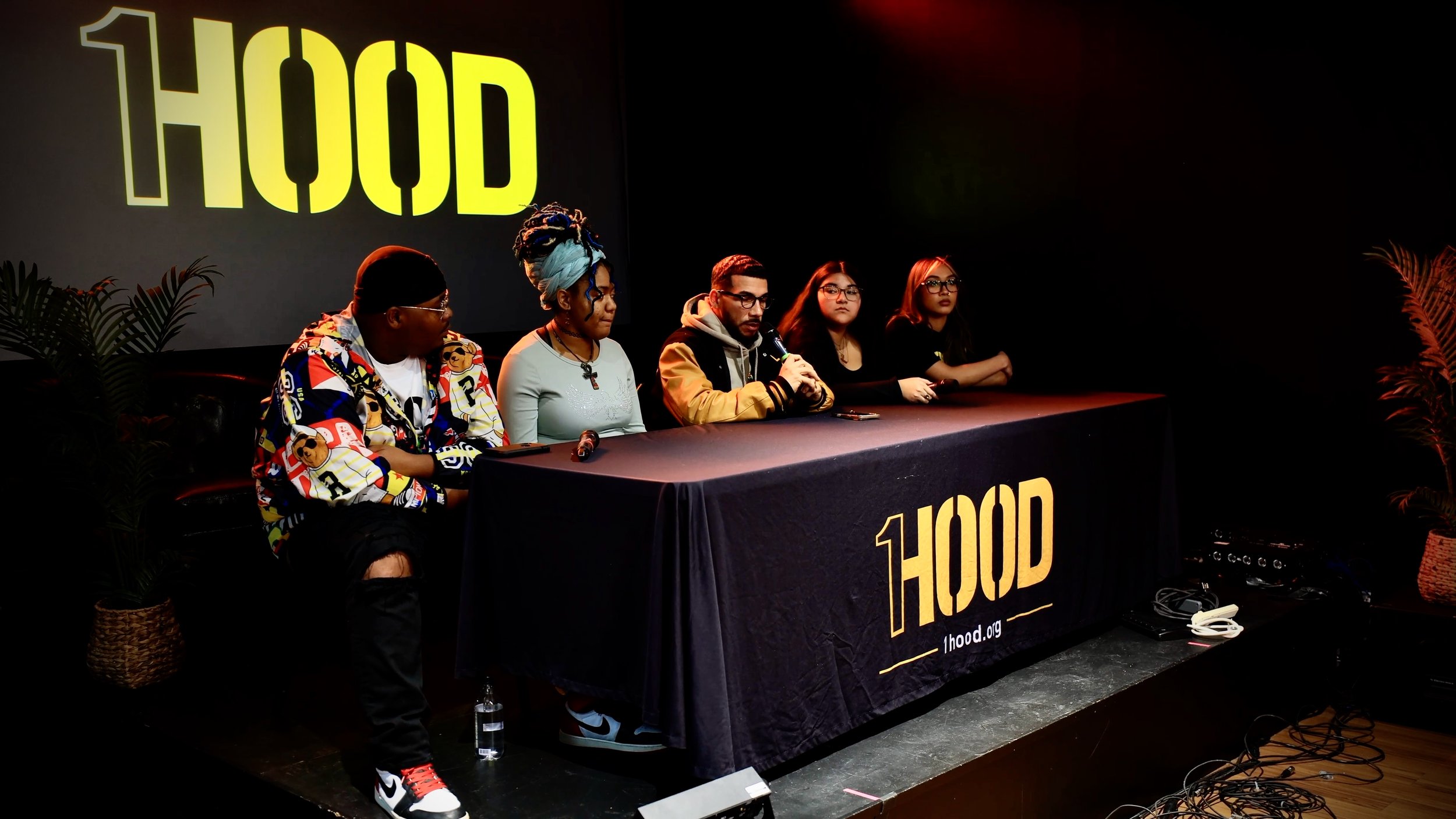 A group of five diverse individuals seated at a table on a stage during a panel discussion or event. Behind them is a large screen displaying the word 'HOOD' in bold yellow letters. The stage is decorated with potted plants on either side, and there are various cables and electronic equipment on the floor.