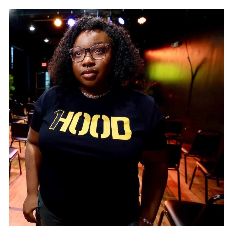 A young woman with glasses and curly hair wearing a black T-shirt with yellow text that reads '1HOOD' standing in a dimly lit room with chairs and colorful lighting.
