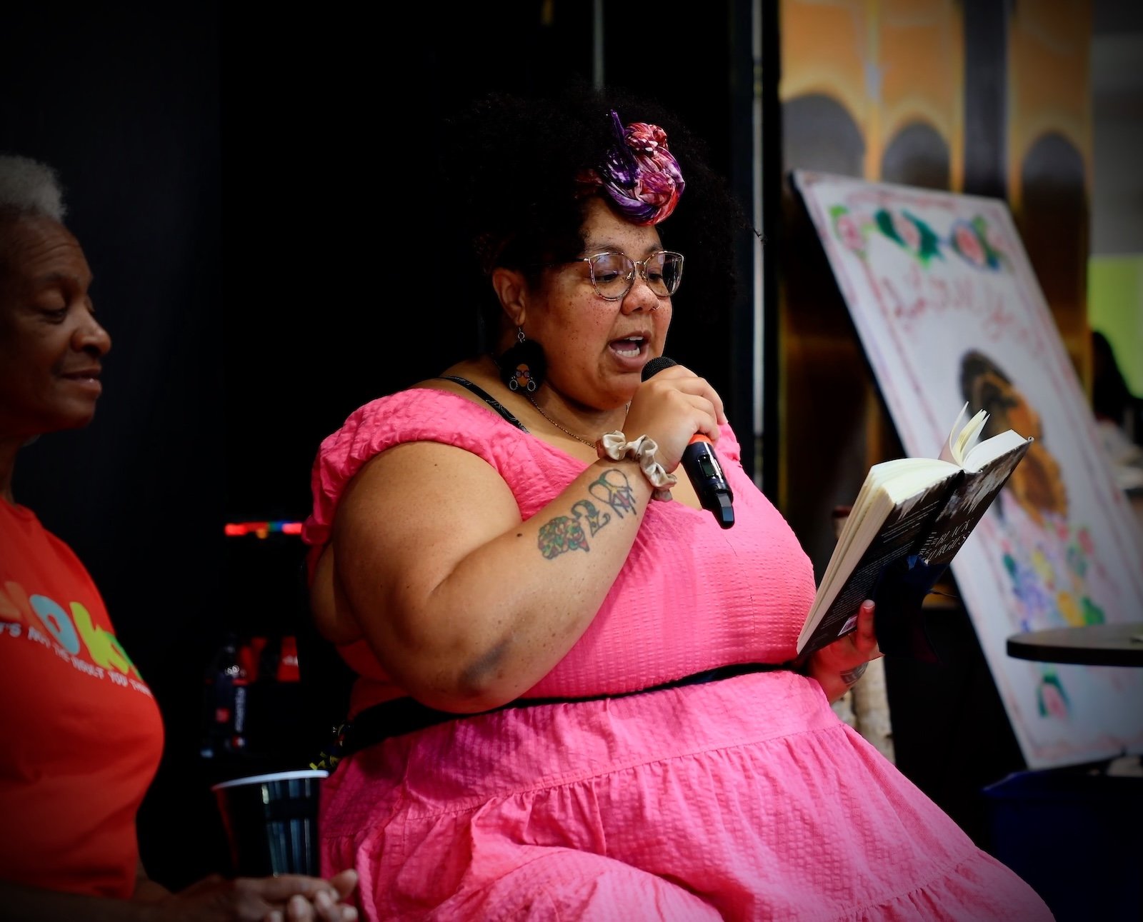 Woman with glasses and curly hair, wearing a pink dress, speaking into a microphone while reading a book. Another woman with short gray hair and a red shirt is partially visible beside her. Background includes artwork on display.
