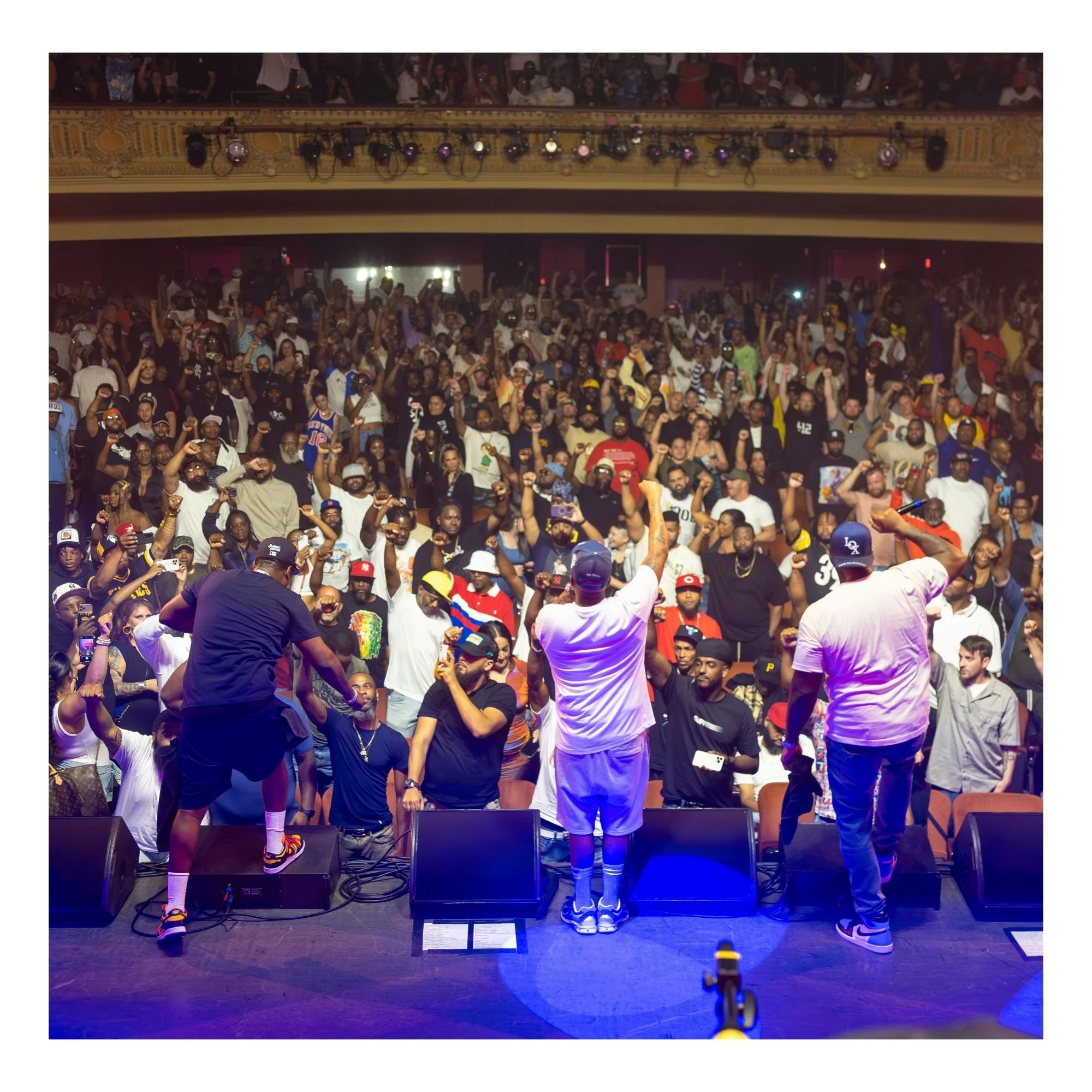 Crowd gathered at a concert or event, facing a stage with performers. Audience members are cheering, some holding drinks or phones, with a balcony above filled with more spectators. Stage lighting casts purple and blue hues.