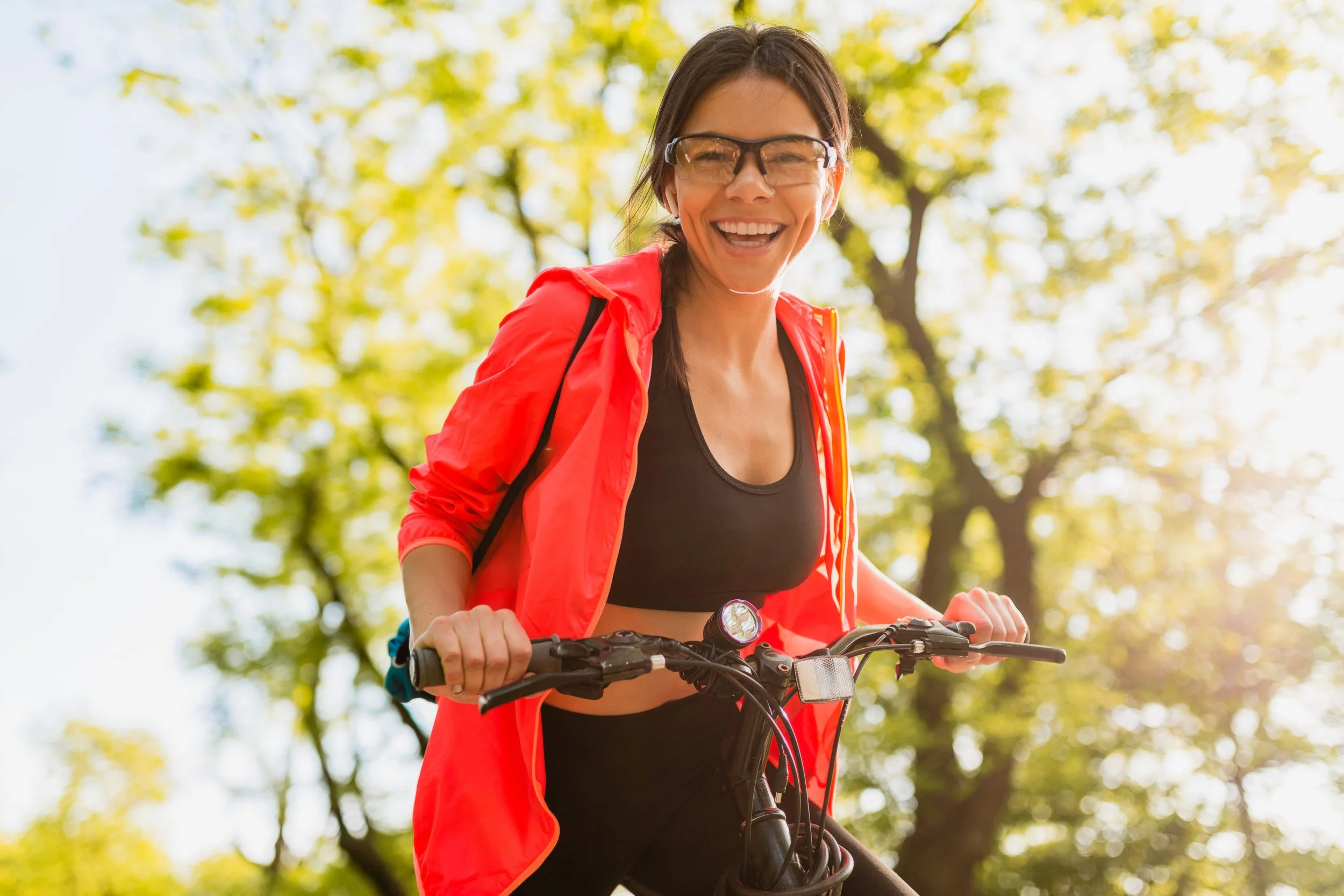A woman smiling while riding a bicycle outdoors during daytime with trees and sunlight in the background.
