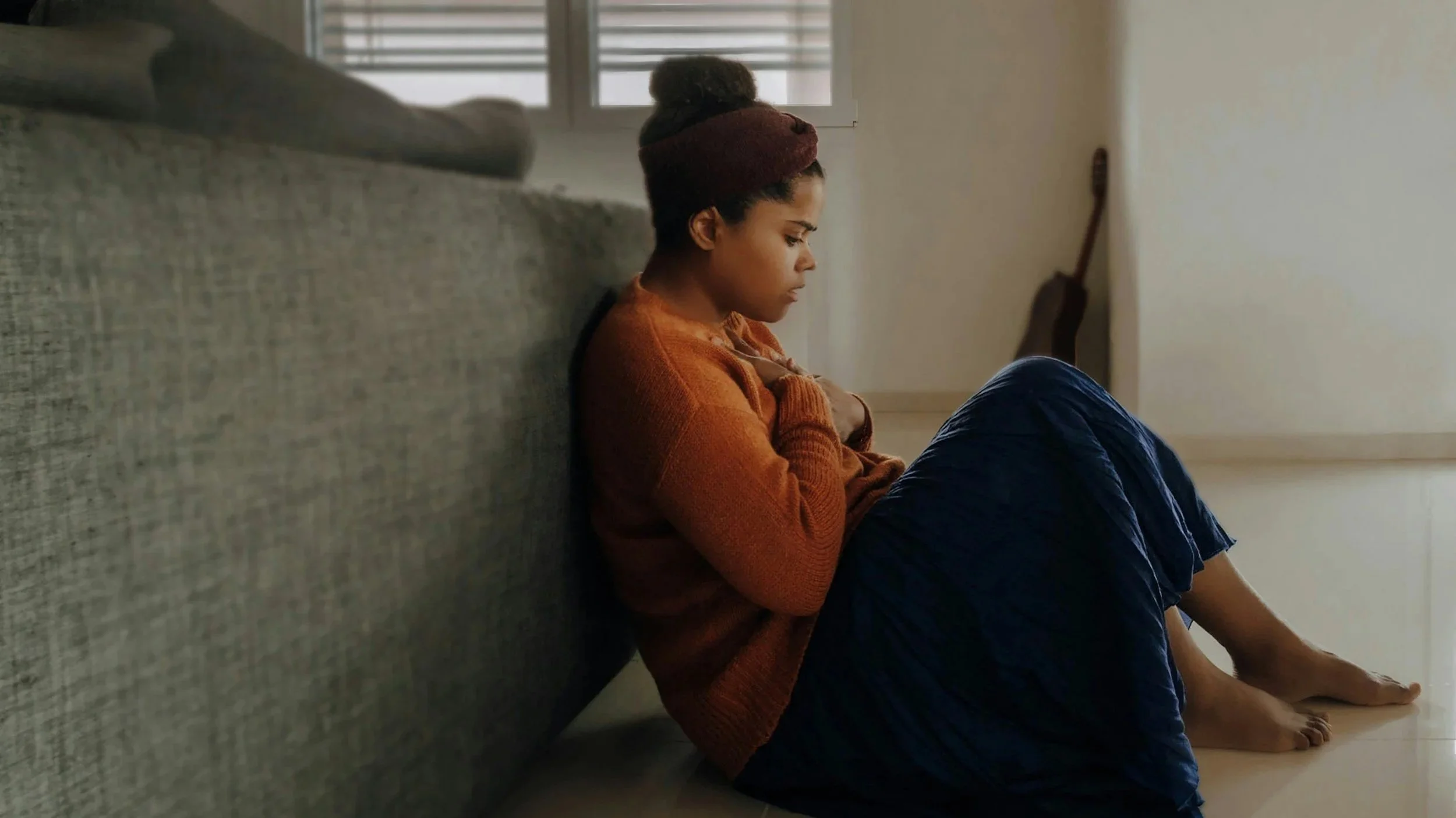 A woman sits on the floor leaning against a gray sofa in a dimly lit room, appearing distressed with her hands held to her chest.