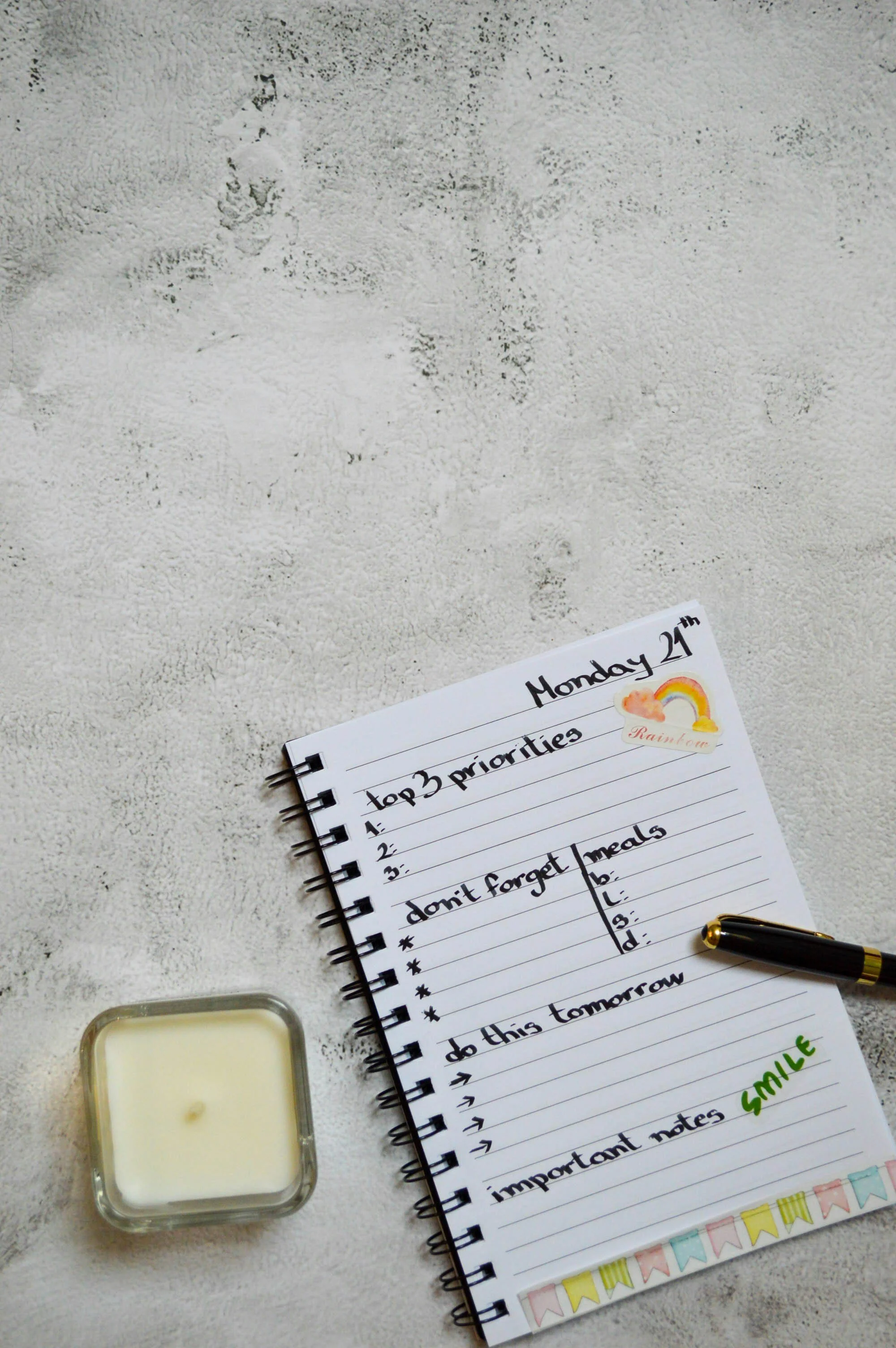 An open spiral notebook with handwritten notes about priorities, important reminders, and to-do list items. The notebook has colorful sticker decorations along the bottom edge. Partially visible are a black pen, a candle, and a small container of cream or lotion on a textured white surface.