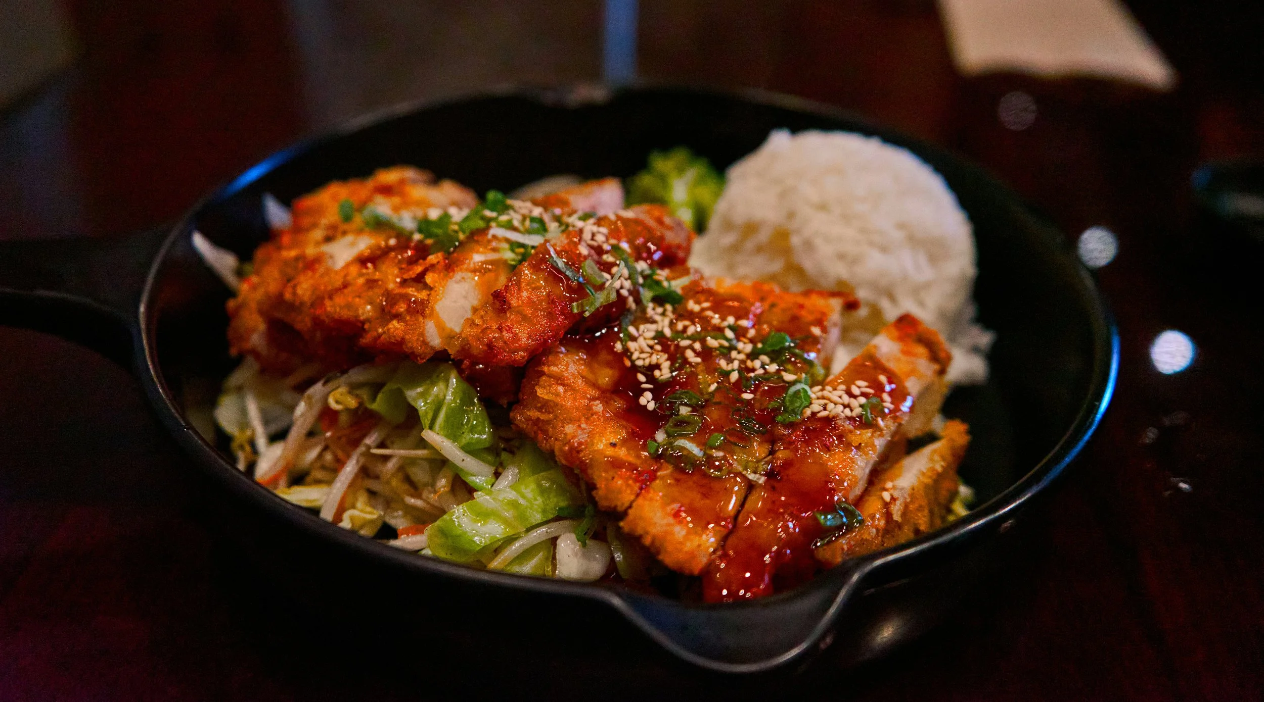 Close-up of a black bowl of Asian-style fish with sauce, green onions, and sesame seeds, served with rice and vegetables.