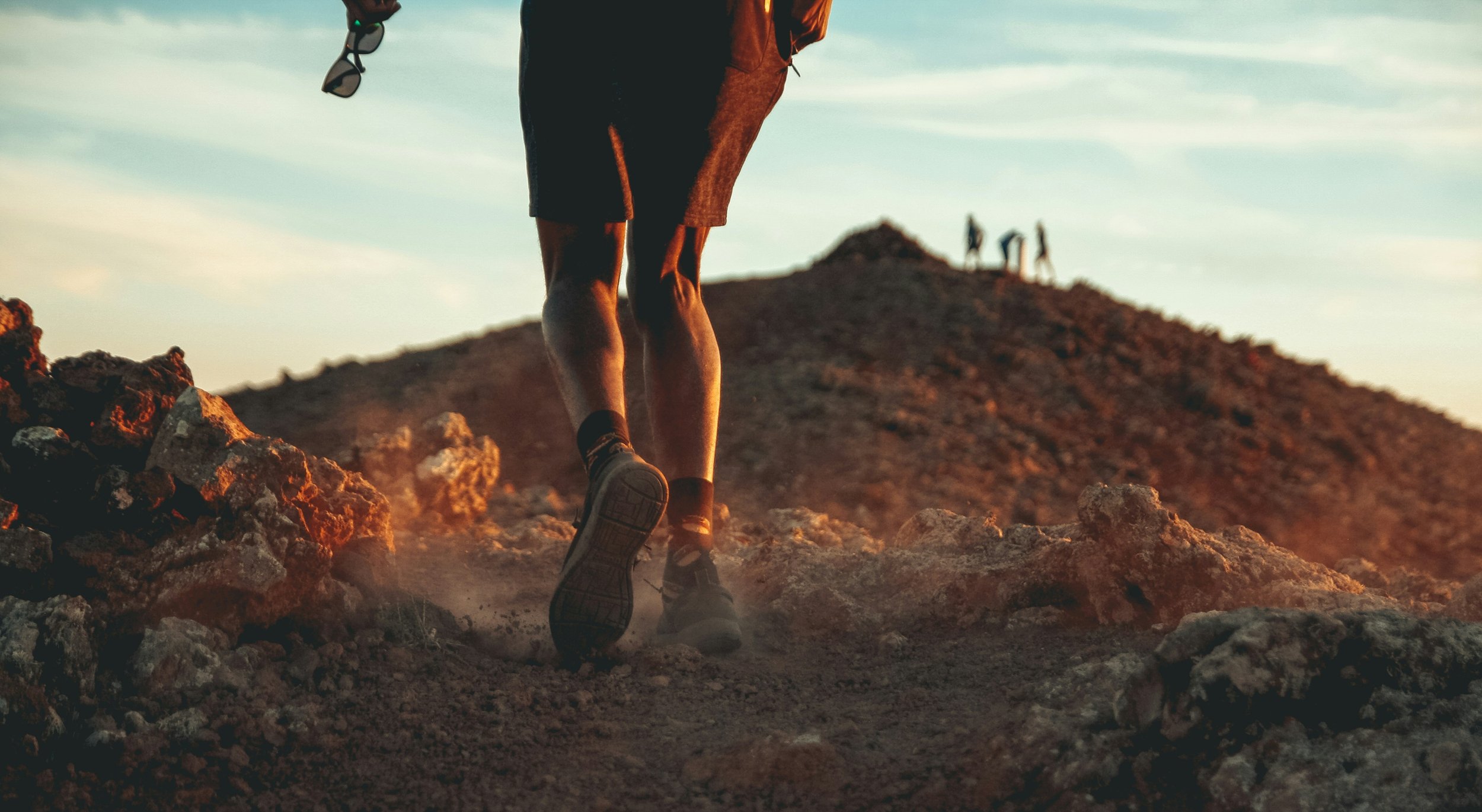 Person hiking on rocky terrain during sunset with a backpack, with three people visible in the distance on a hilltop.