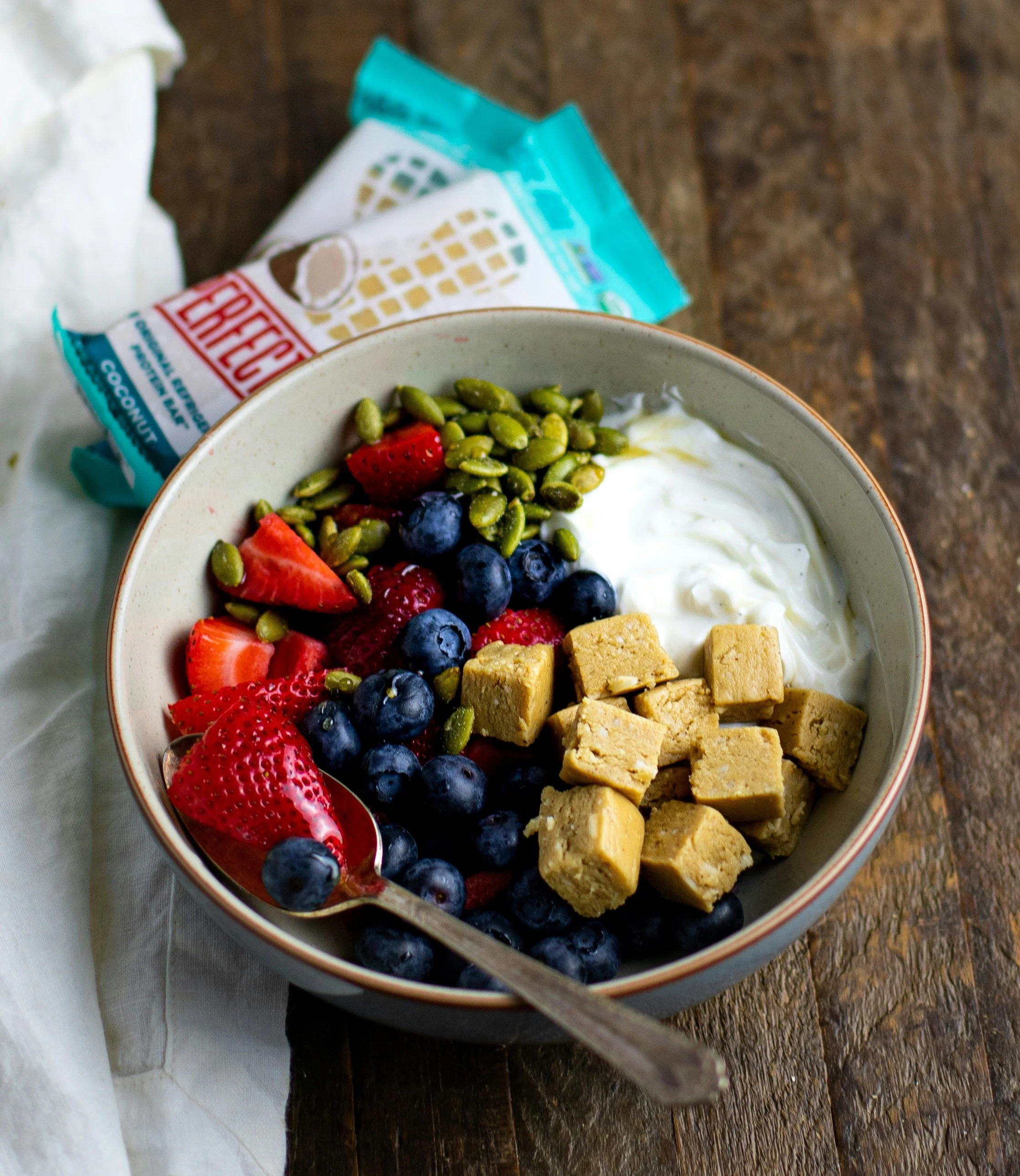 A bowl of yogurt topped with strawberries, blueberries, pumpkin seeds, and small cubes of white or yellowish cereal or granola. In the background, there is a package of frozen strawberries and a white napkin on a wooden surface.