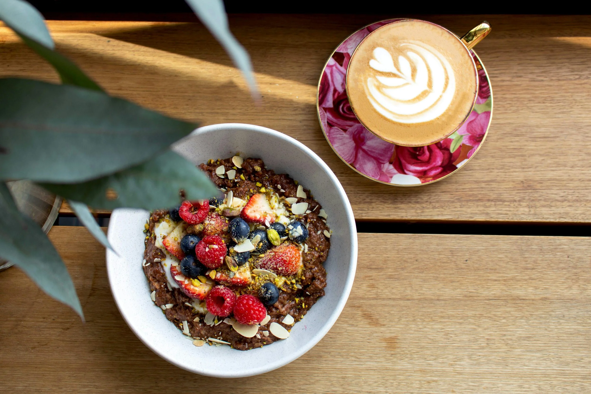 Overhead view of a cup of coffee with latte art on a floral saucer, and a bowl of oatmeal topped with fruits and nuts on a wooden surface, partially obscured by green leaves.