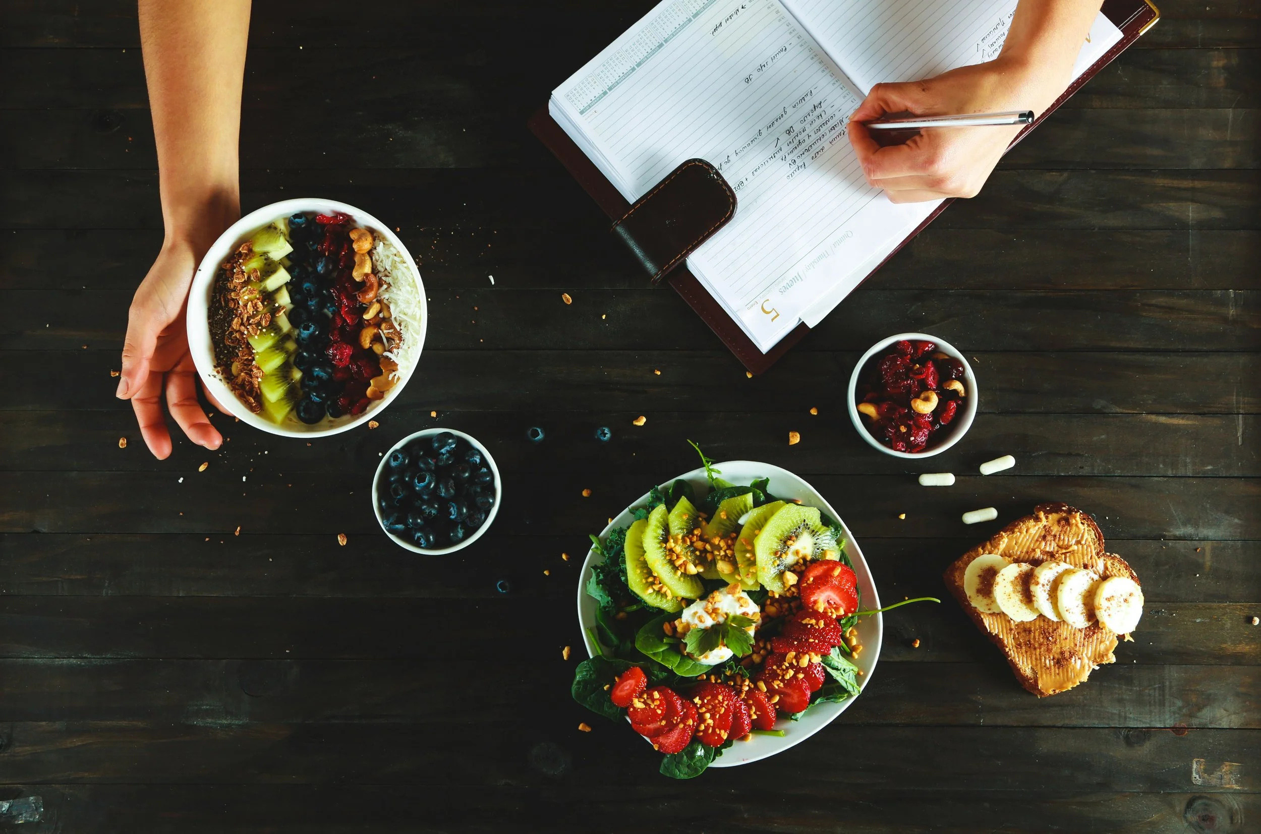 Overhead view of a table with bowls of fruit and a person writing in a planner.