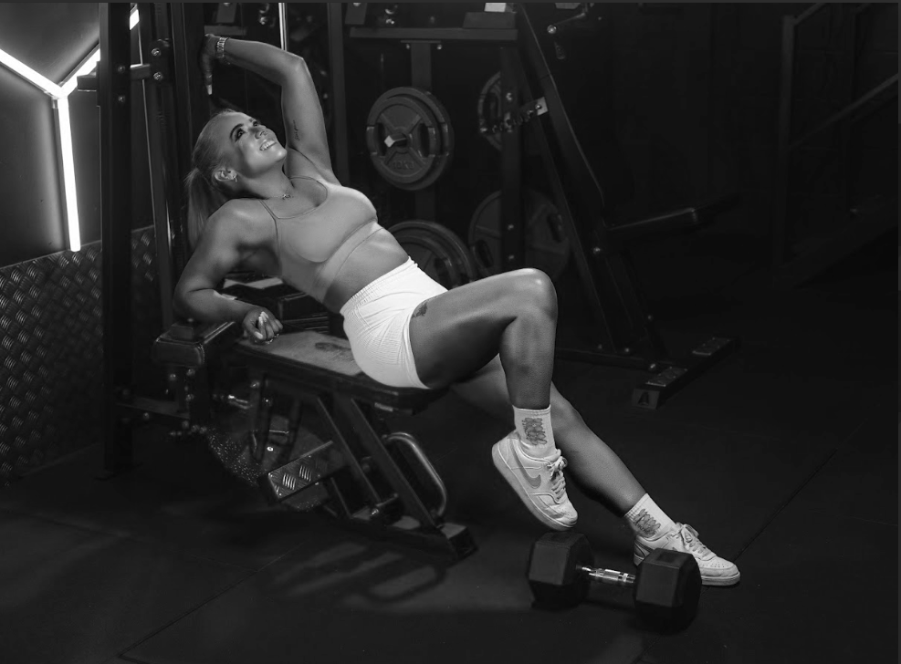 A woman lifting a dumbbell in a gym, sitting on a workout bench, smiling, with weight plates in the background.