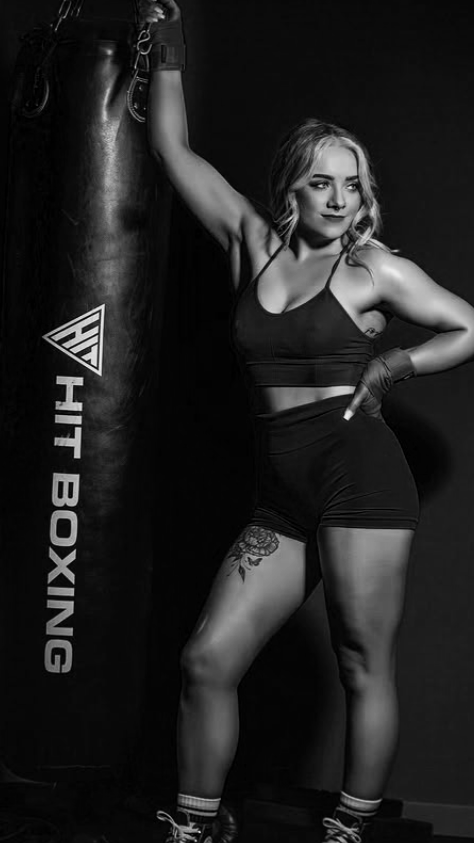A woman in athletic attire leans against a hanging punching bag with her left arm, in a boxing gym. She has a tattoo on her thigh and wears boxing gloves and athletic shoes.