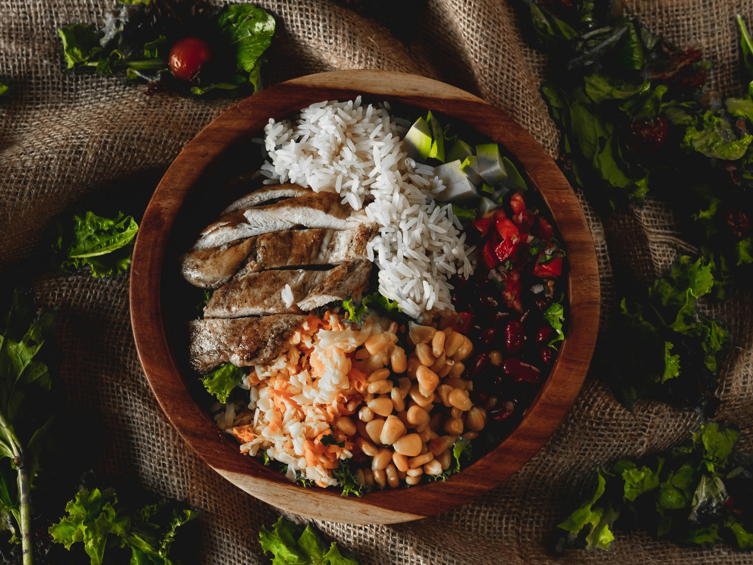 A wooden bowl filled with cooked white rice, grilled fish, and chopped vegetables, on a textured burlap cloth surrounded by fresh greens and herbs.