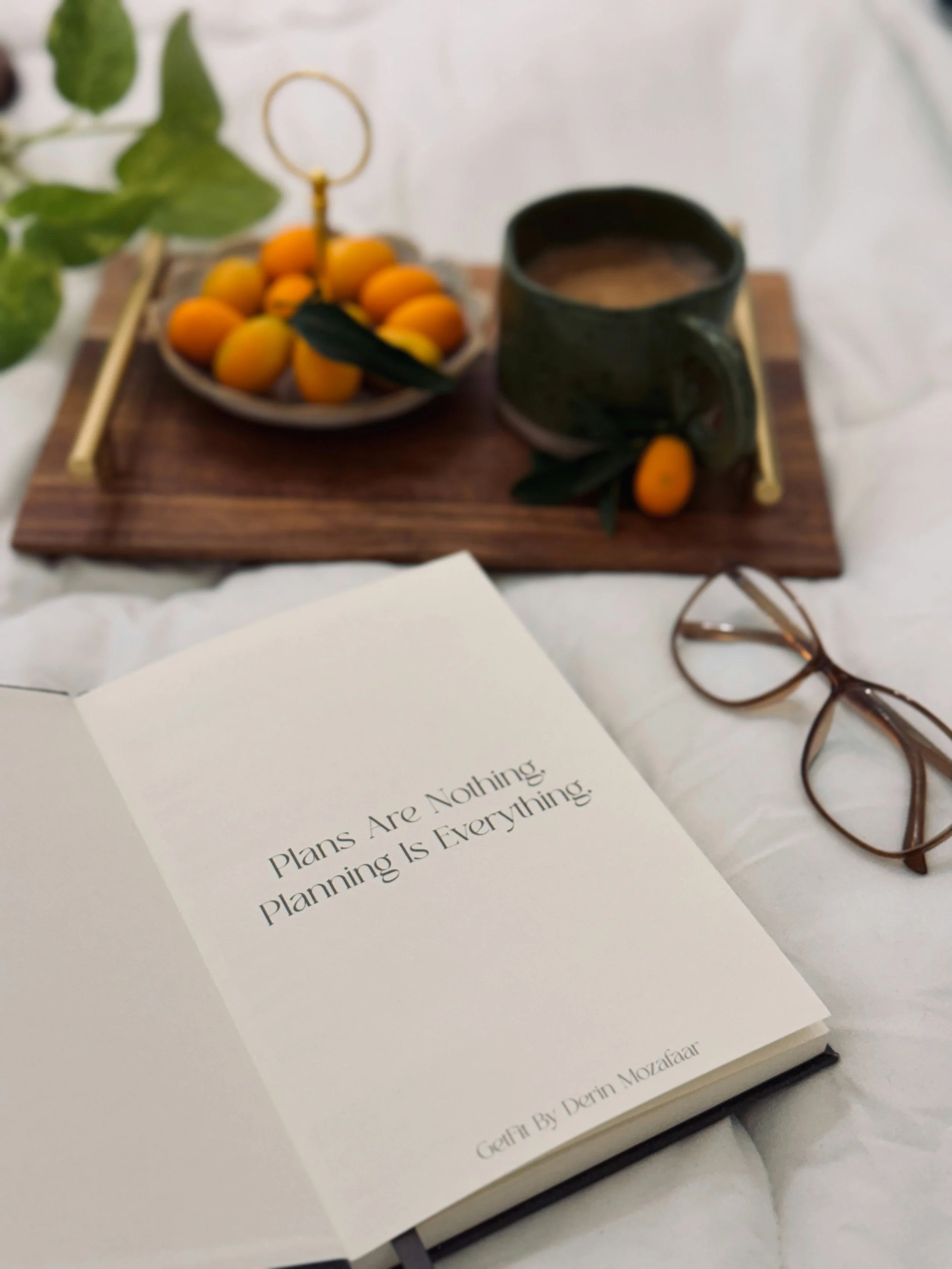An open notebook with the quote 'Plans Are Nothing, Planning Is Everything' by Dan Mozaffar on the page, a pair of glasses, and a white table with a mug, a plate of small orange fruits, and some green leaves in the background.