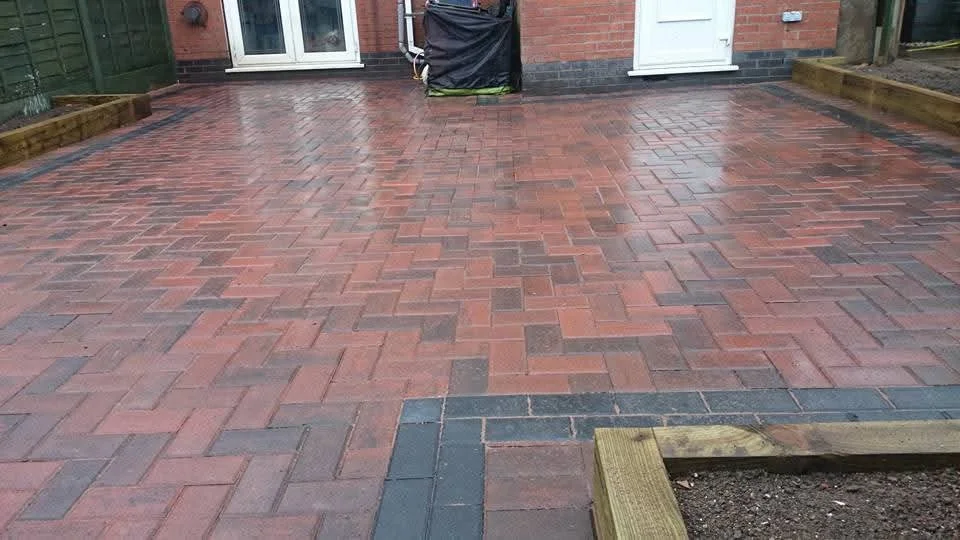 Newly installed red and black brick paver patio with a small section of unfinished border in the foreground, in a backyard with a brick house and white door in the background.