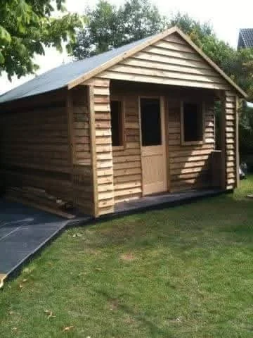 Wooden garden shed with a sloped roof, a door, and two windows, situated on a grassy yard with trees in the background.