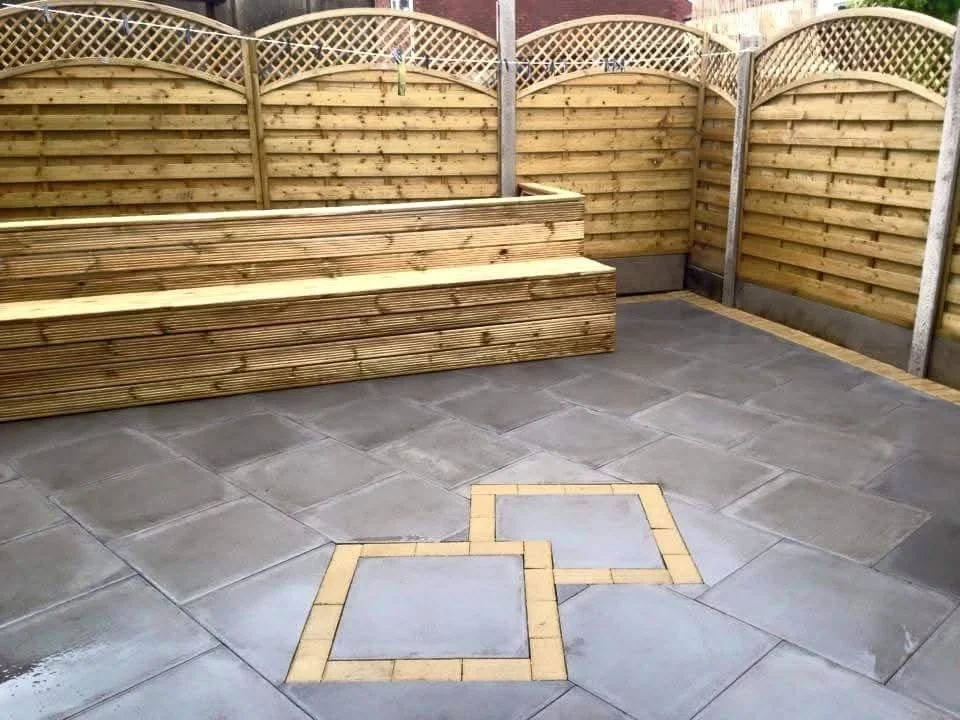 Empty backyard patio with gray paving stones, a wooden fence, and two square decorative tiles in the center.