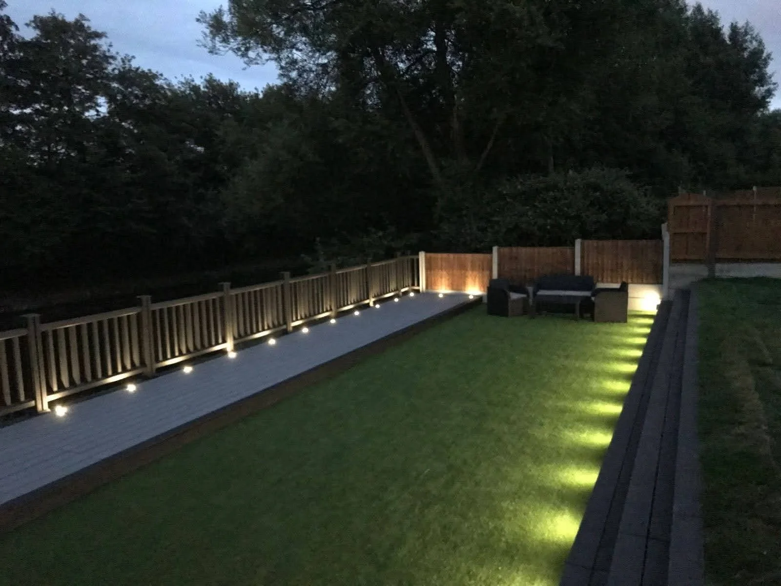 Nighttime backyard with a wooden deck, outdoor seating, and low-level landscape lighting illuminating the patio area and surrounding grass.