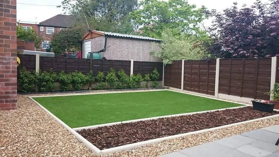 A backyard with a small green lawn, bordered by white edging, surrounded by gravel and brown mulch, with a wooden fence and trees in the background.