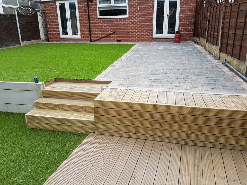 Backyard with a wooden deck, artificial grass, and brick patio with a house in the background.