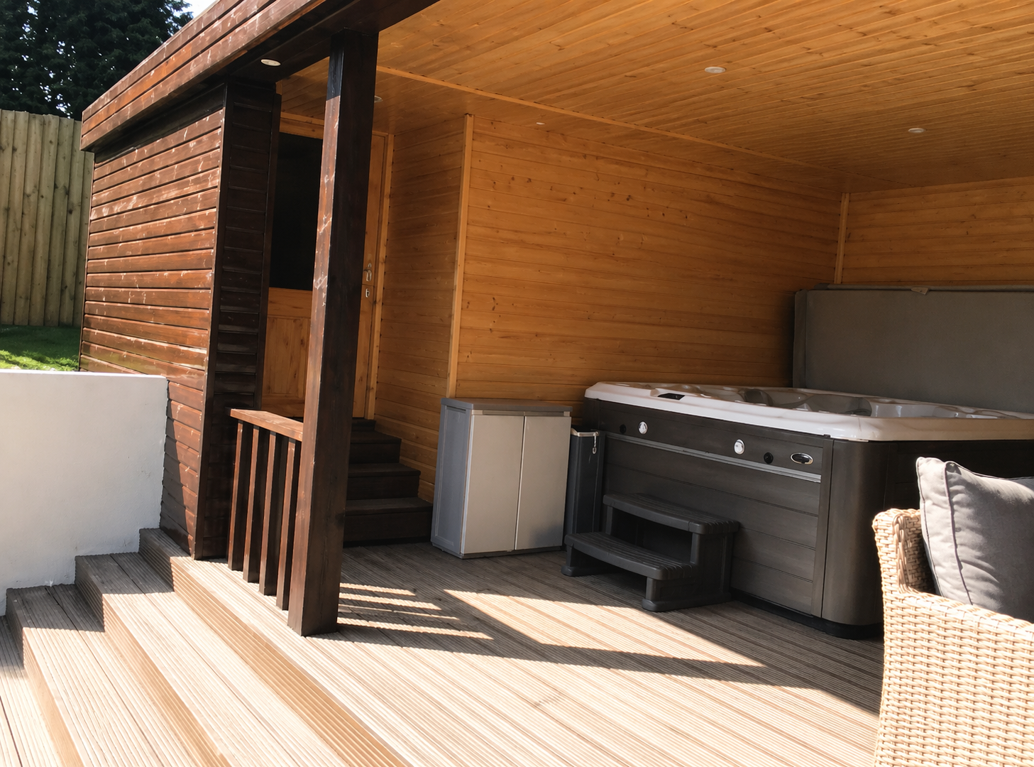 Outdoor wooden deck with hot tub, small fridge, and cushioned chair, part of a wooden patio area with steps leading down and a wooden privacy fence in the background.