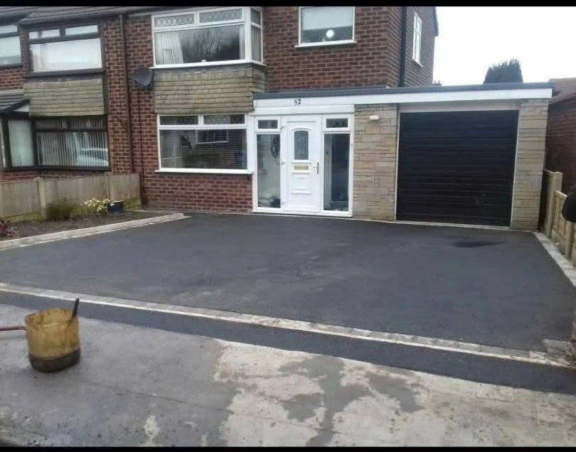 Front view of a house with a freshly paved driveway, white front door, and an attached garage with a black door. There is a wooden fence on the right and some flower beds on the left.
