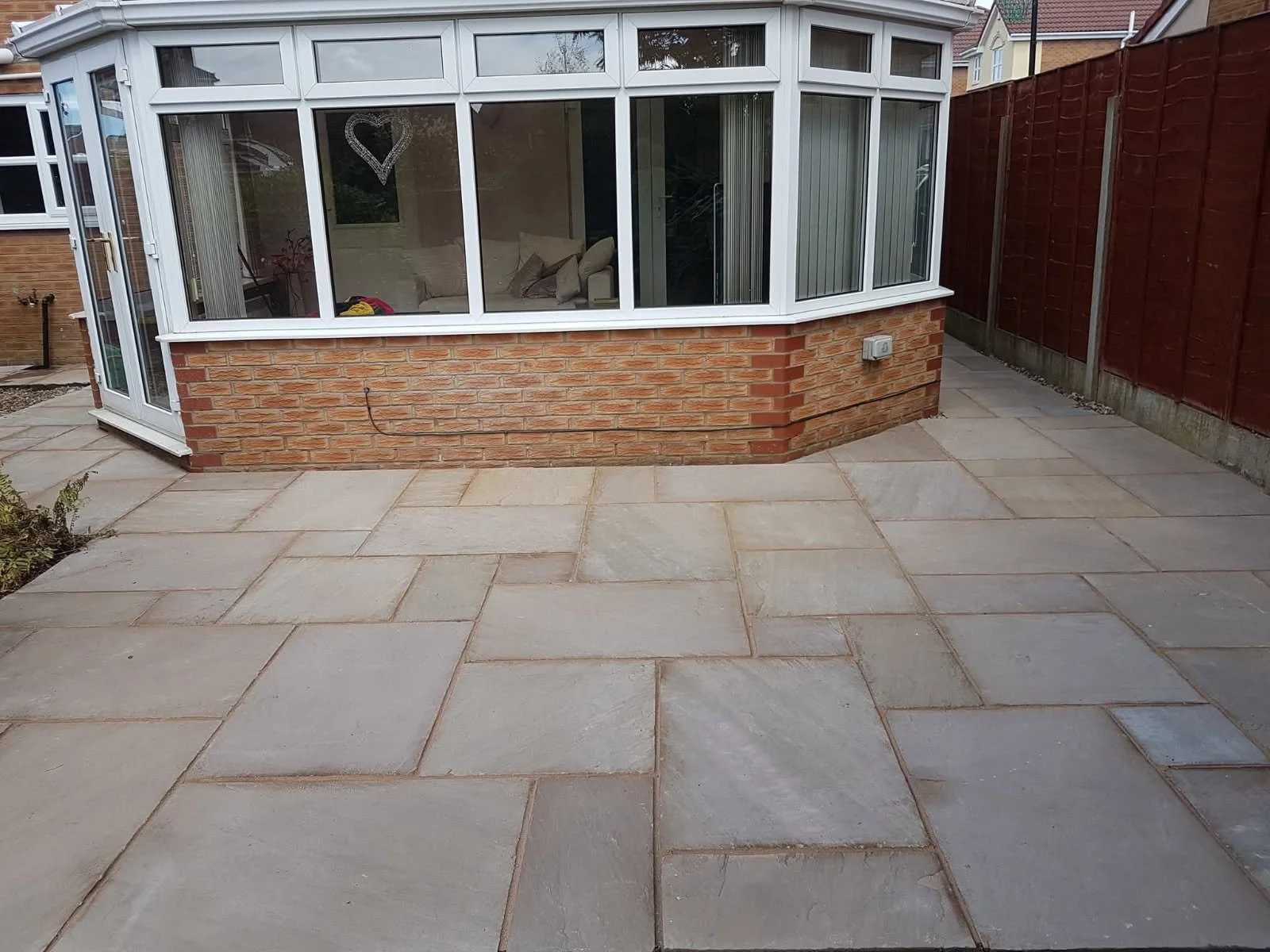 A backyard patio with large rectangular stone pavers, a brick wall with a white framed conservatory, wooden fencing on the right, and a small garden area on the left.