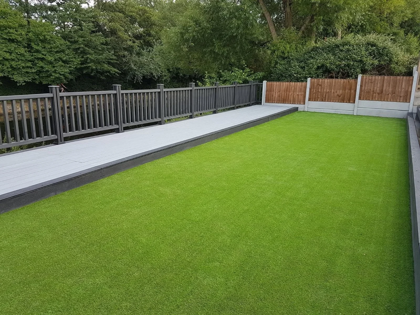 A backyard with a bright green artificial grass lawn, a white wooden deck, and a black wooden fence, with trees and shrubbery in the background.