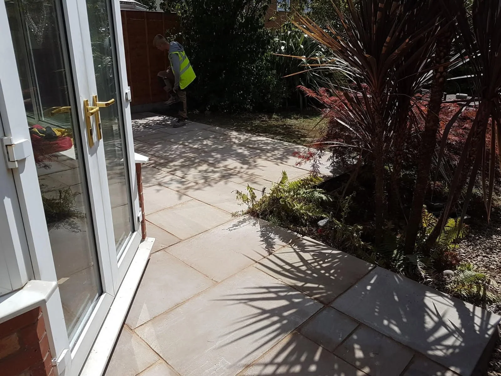 View of a backyard patio with a person in a yellow safety vest sitting on a wooden bench near the fence, surrounded by trees and plants, sunlight casting shadows on the ground.