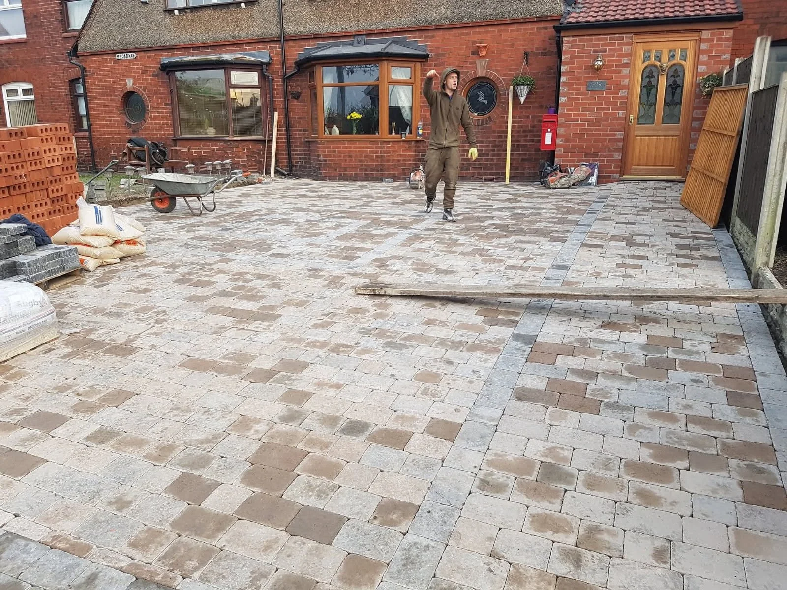 A man working on laying a brick patio in front of a red brick house. Construction materials and tools are scattered around, including a wheelbarrow, bags of mortar, and bricks. The man is wearing work clothes and gloves.