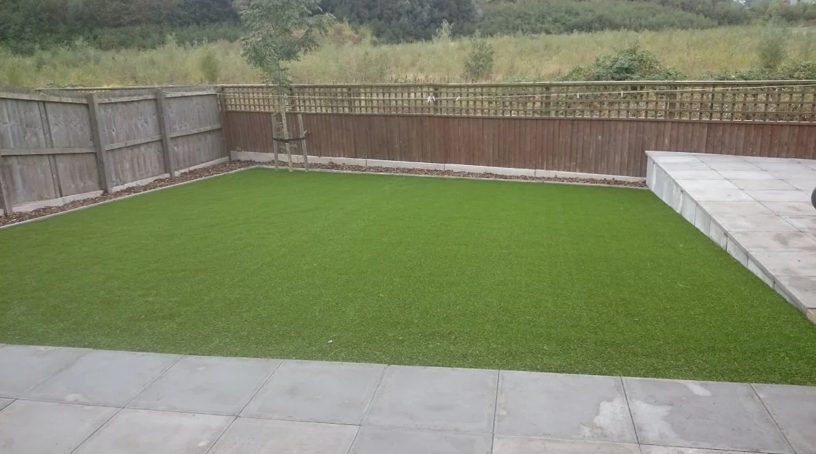 A backyard with a concrete patio, artificial grass, a wooden fence, and a small tree in a pot.