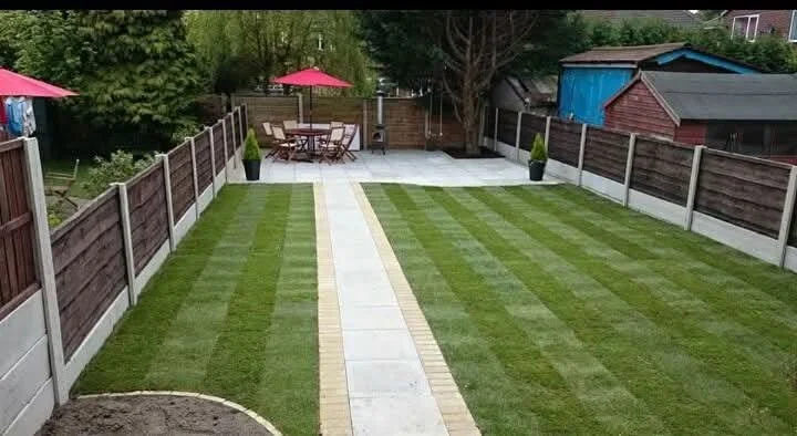 View of a backyard with a paved walkway running through a lawn with striped grass patterns, fenced sides, and outdoor furniture and umbrellas at the far end.