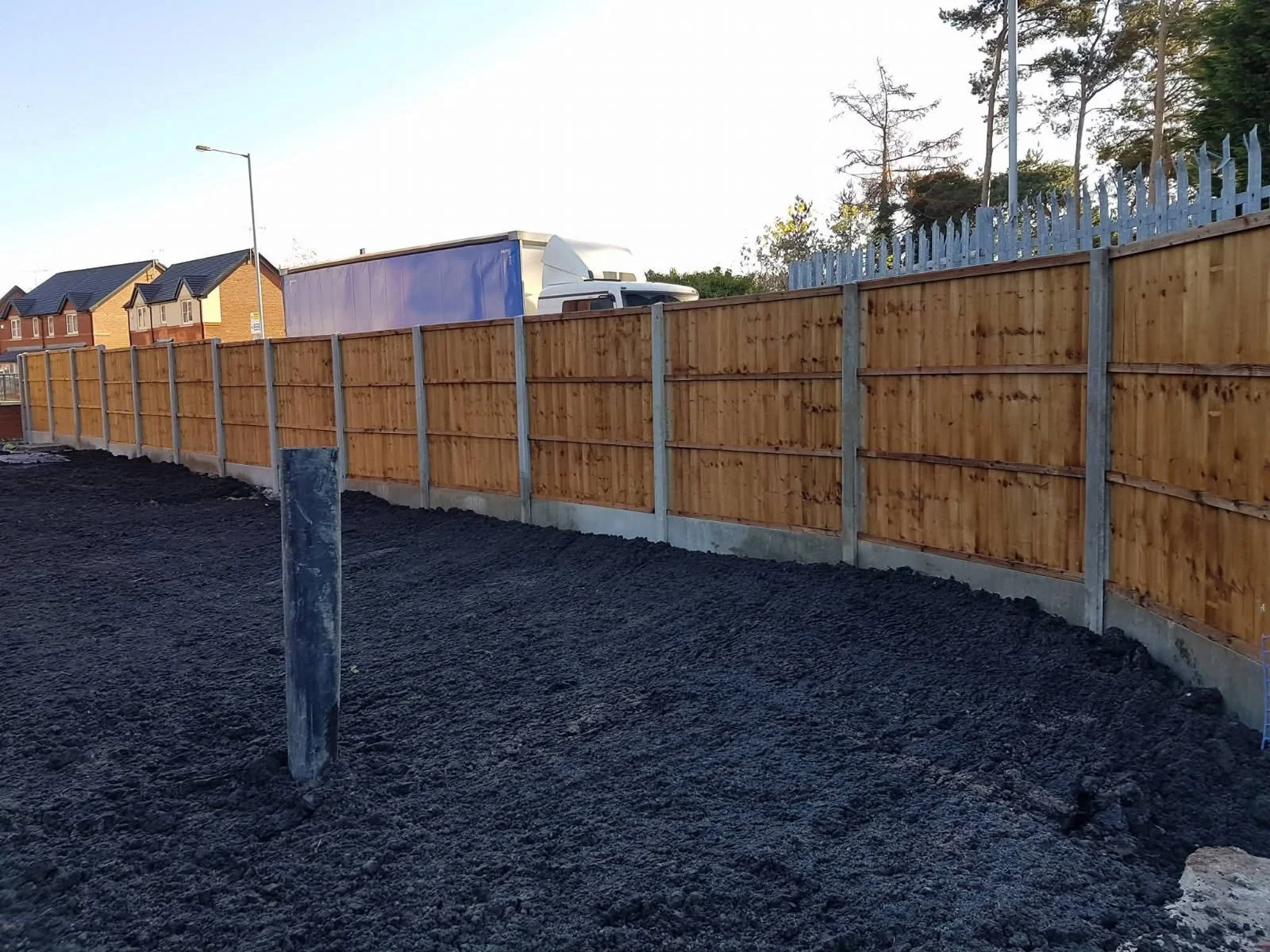 Newly constructed wooden fence along a dark soil ground in a residential neighborhood with houses and trees in the background.