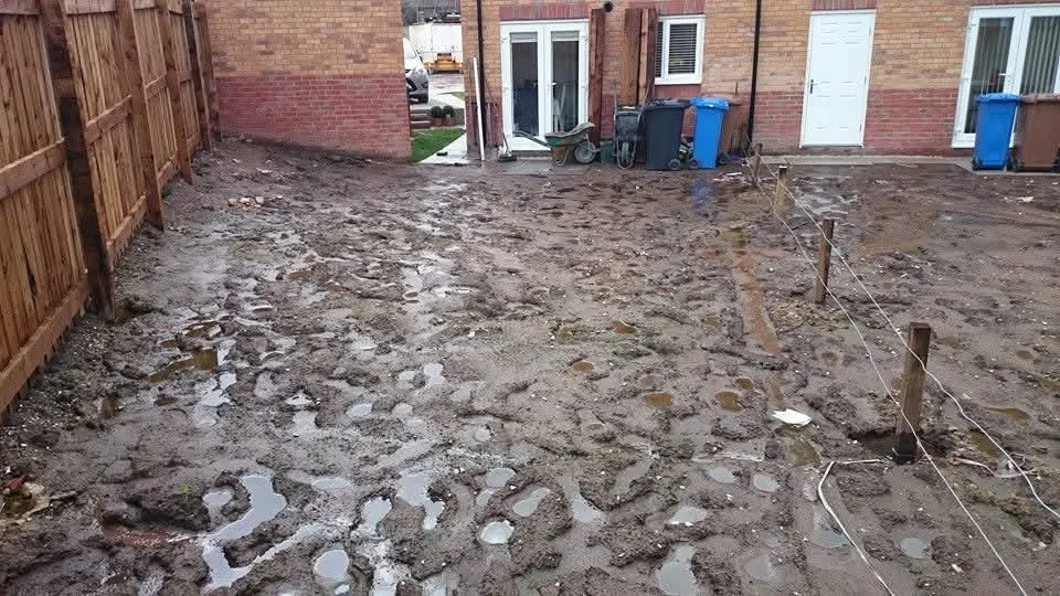 A backyard with muddy, uneven ground and wooden stakes with string marking a boundary, fences on the sides, and houses in the background.