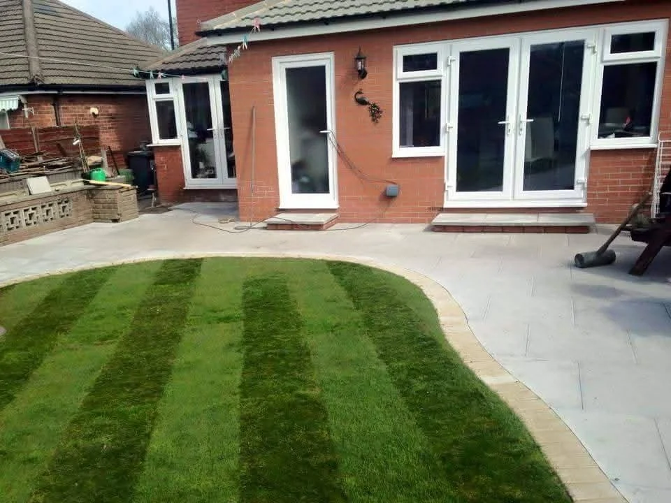 Backyard with a small, striped green lawn and stone patio. The patio is adjacent to a red brick house with white-framed double glass doors and windows.