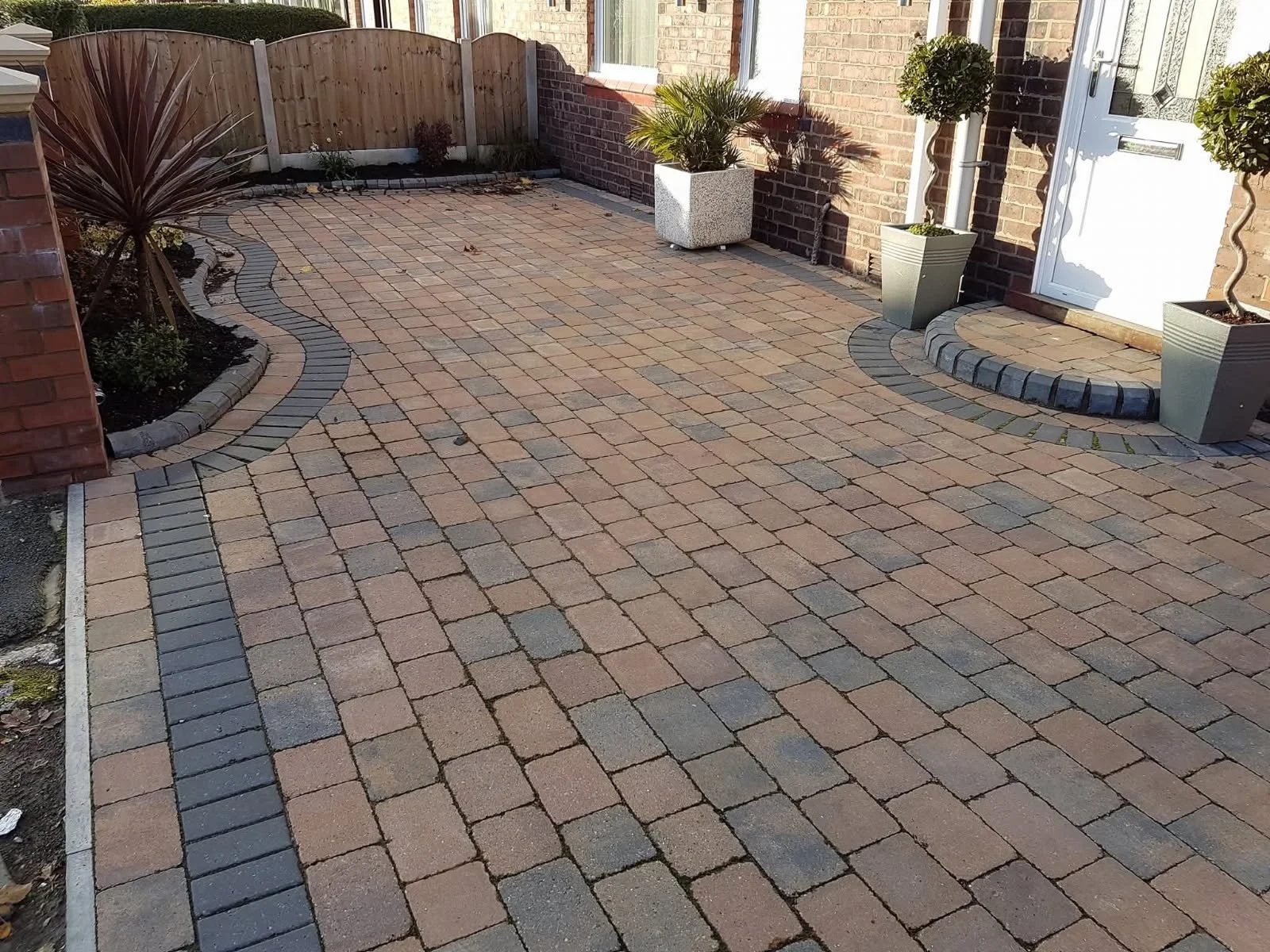 Paved front yard with brick and stone pavers, curved border, and potted plants near the house entrance.