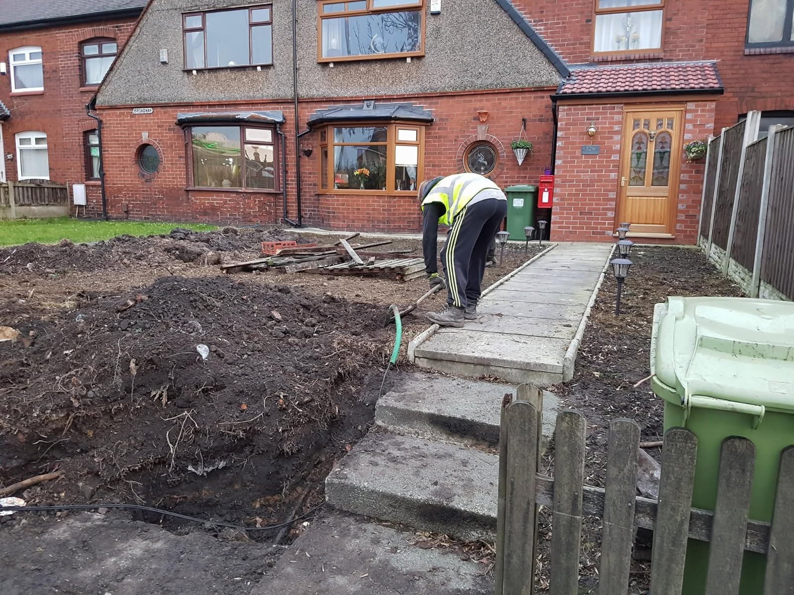 A person working on laying a concrete pathway in the front yard of a house. The person is wearing a high-visibility vest and bending over as they work. The yard is currently dug up, with dirt and construction materials visible. The house is a brick building with a wooden front door, some garden decorations, and a small path leading to the entrance.