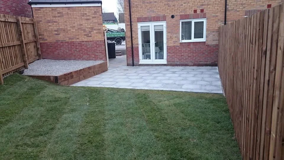 Backyard with a grassy area leading to a paved patio, enclosed by wooden fencing, in front of a brick house with white doors and windows.