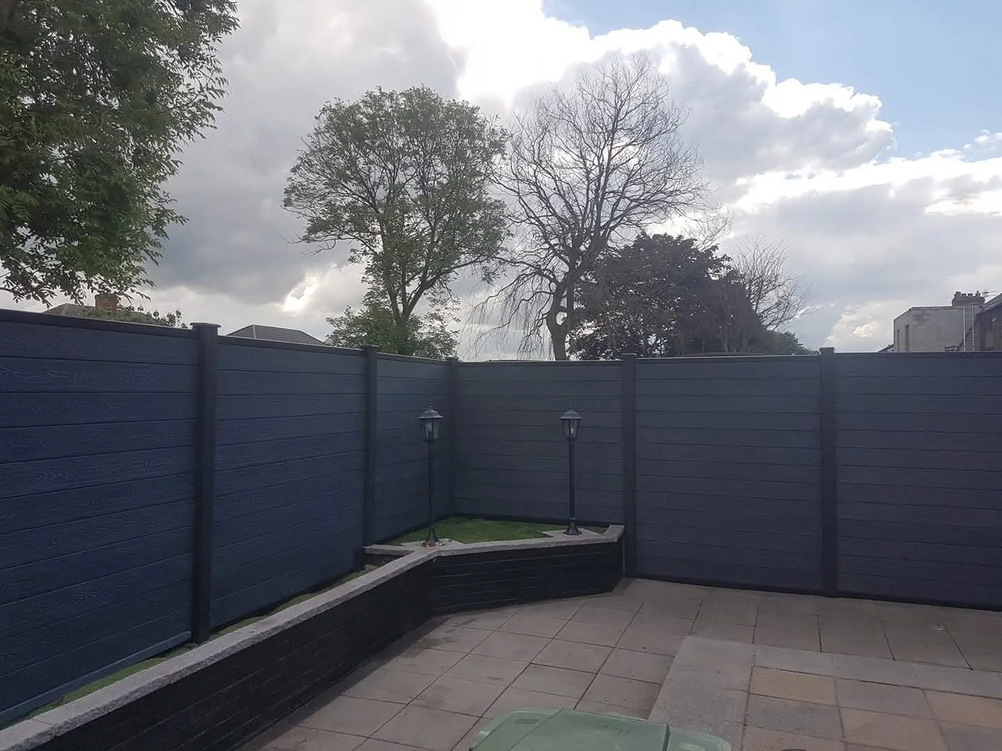 A backyard patio with gray tiles, a dark blue wooden fence, and a small grassy area with two black lamp posts. There are trees and a cloudy sky in the background.