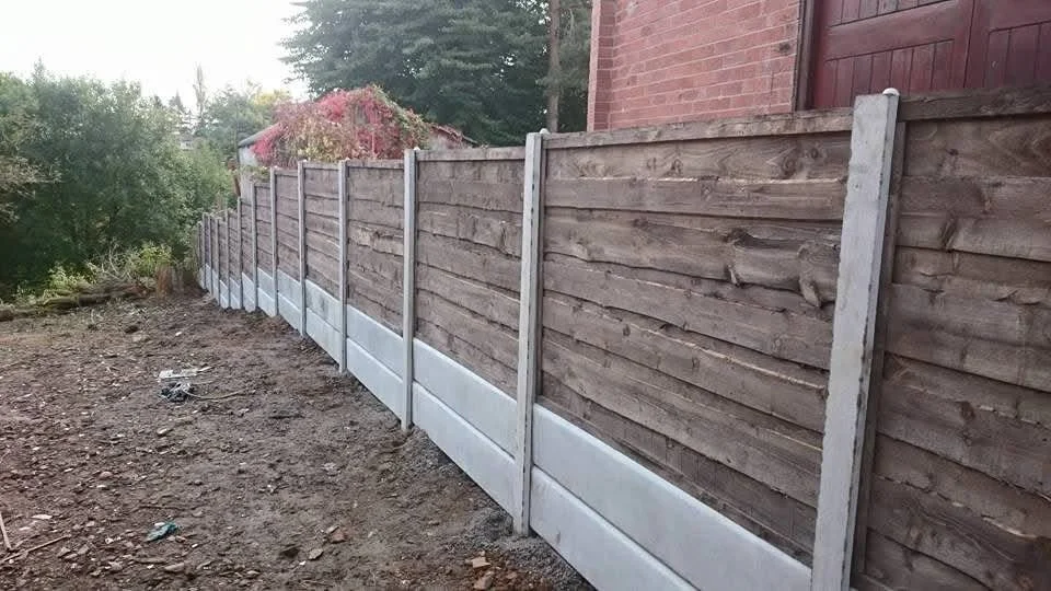 Newly constructed wooden fence with concrete base in a backyard, with soil and gravel in the foreground and trees and a red brick building in the background.