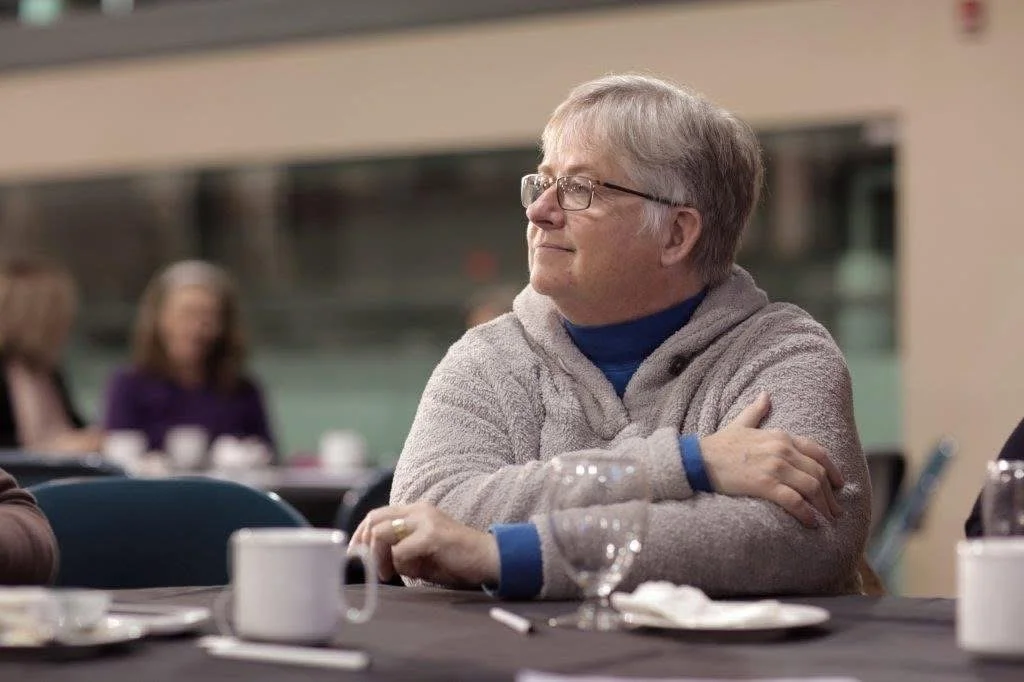 Marianne sits at a table, looking into the distance with a small smile.