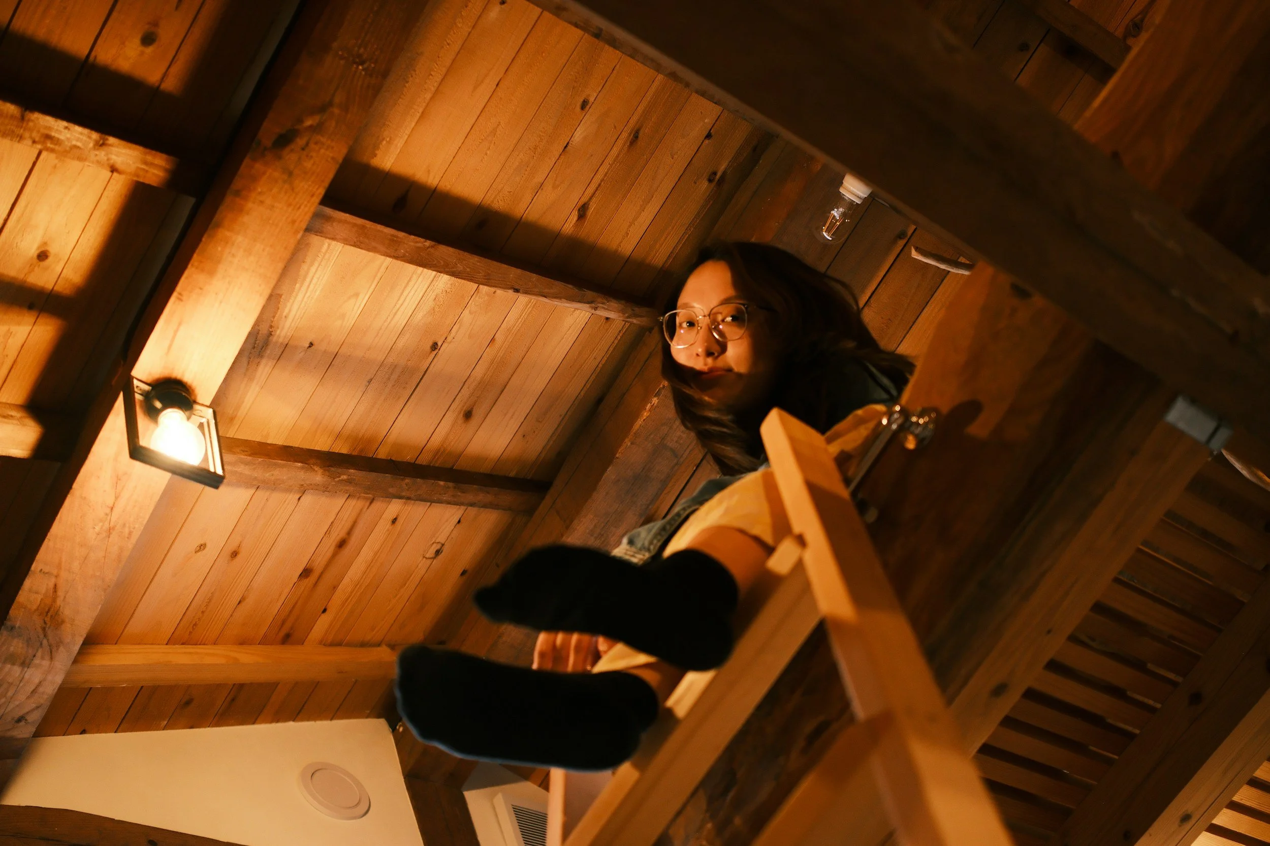 A woman with glasses and dark hair looking down from a wooden loft or balcony, surrounded by wooden beams and ceiling panels in a rustic setting, with a warm light fixture overhead.