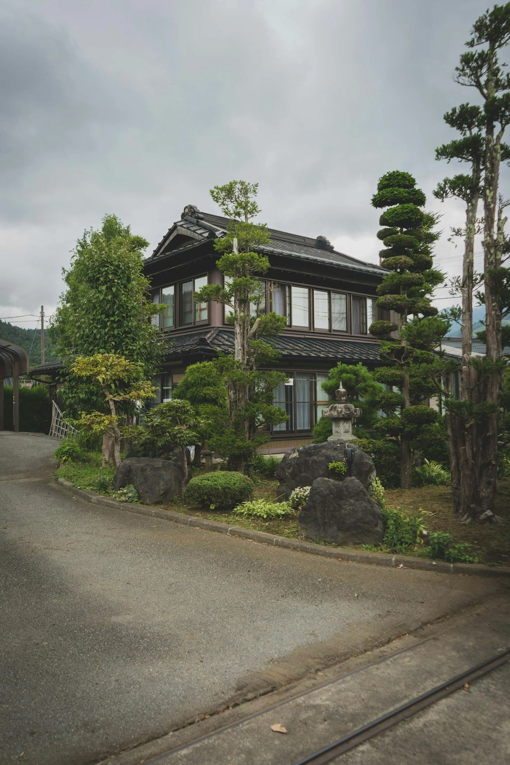 Traditional Japanese house with dark wooden exterior, surrounded by well-manicured trees and landscaping, cloudy sky overhead.