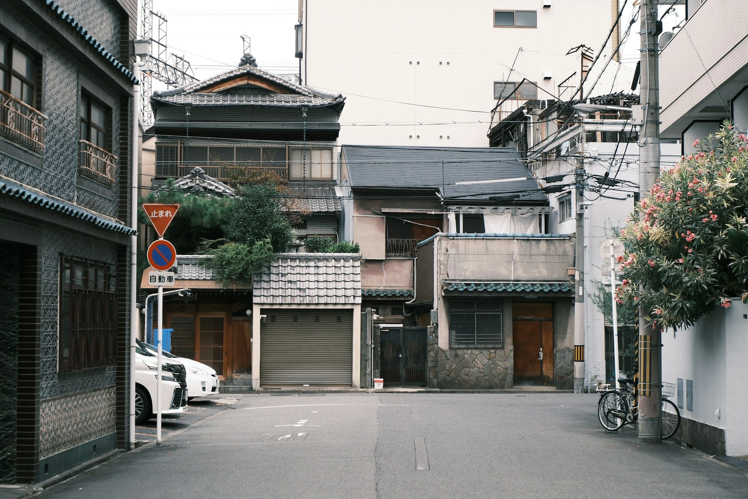 A quiet urban street scene in Japan with traditional and modern buildings, parked cars, bicycles, utility poles, and street signs.