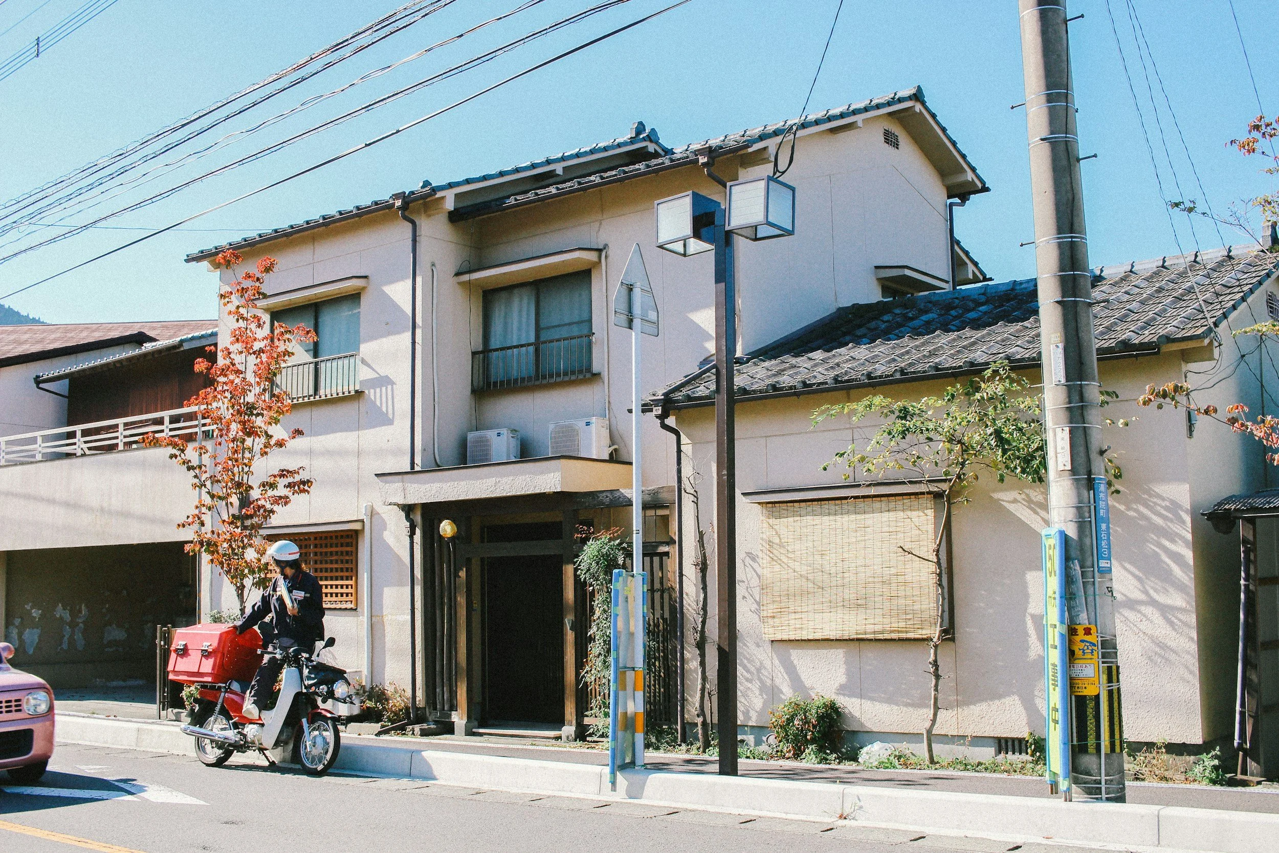 A street scene with a two-story house, a person riding a scooter with a red delivery box, and a utility pole, under a clear blue sky.