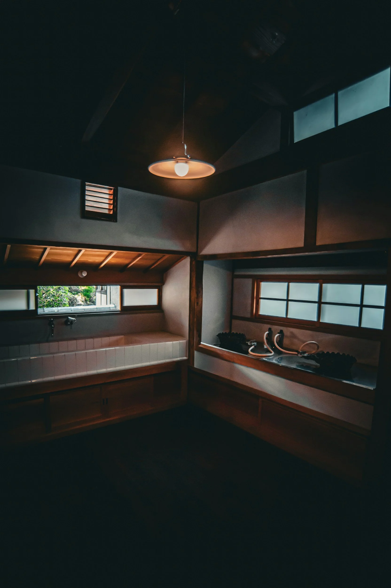 Interior of a traditional Japanese room with ceiling light, wooden beams, sliding windows, and a small bathing area with a pink tiled tub and taps.
