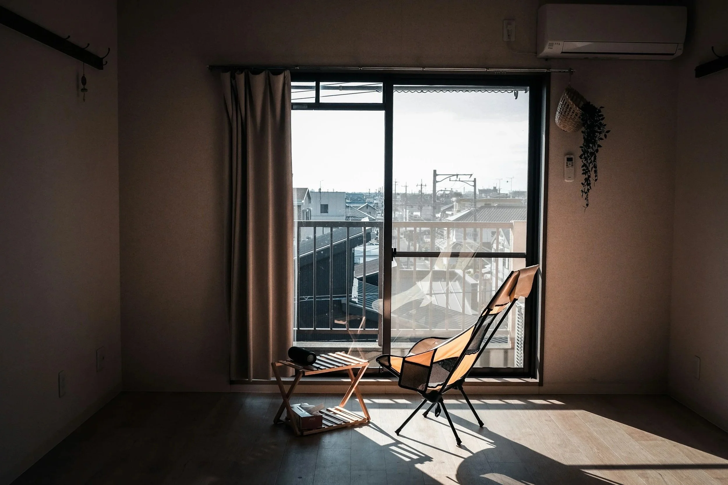 Sunlit interior room with a large window, beige curtain, and a balcony. There is a beige lounge chair, a small wooden table, a black speaker, and a few books inside the room. An air conditioning unit is mounted on the wall.