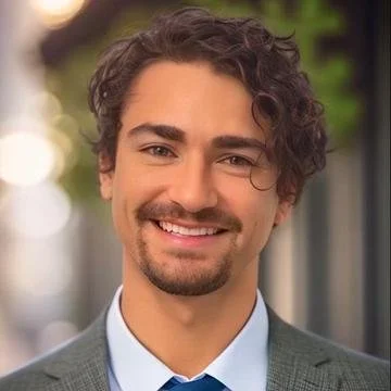 A young man with curly hair and a beard smiling outdoors.