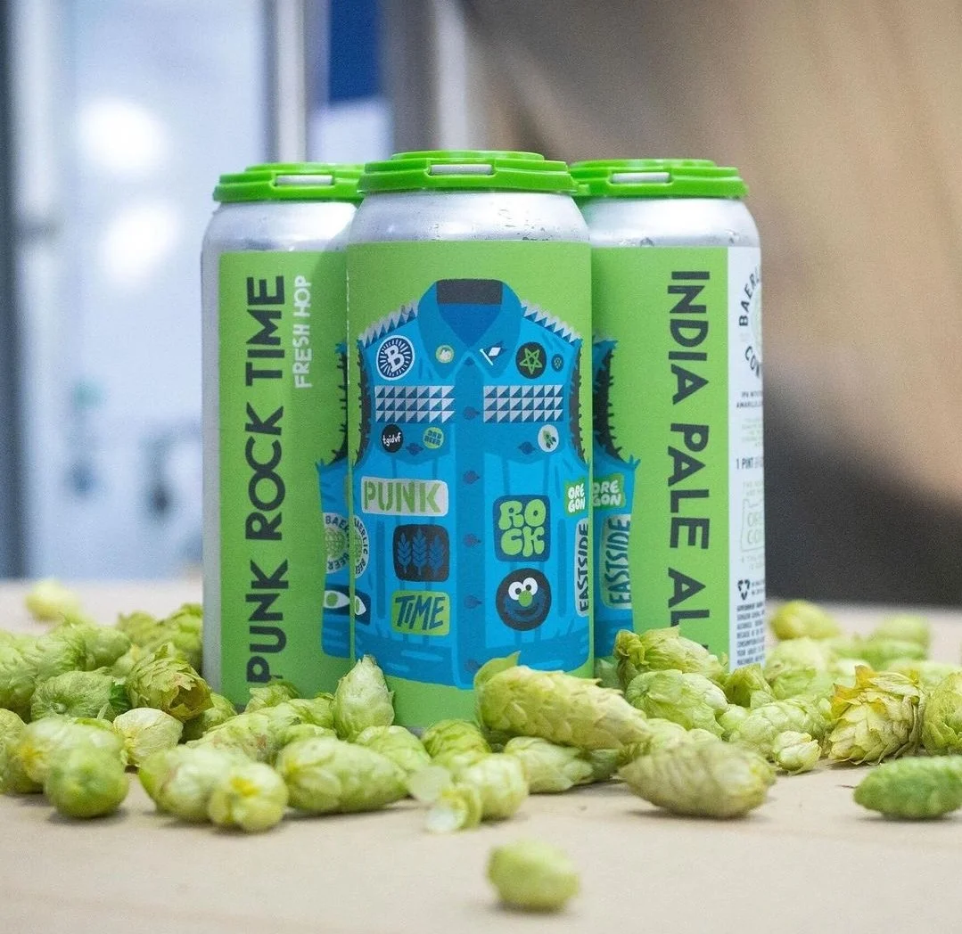 Cans of "Punk Rock Time Fresh Hop" India Pale Ale with green labels and cartoon vest design, surrounded by fresh hops on a table.