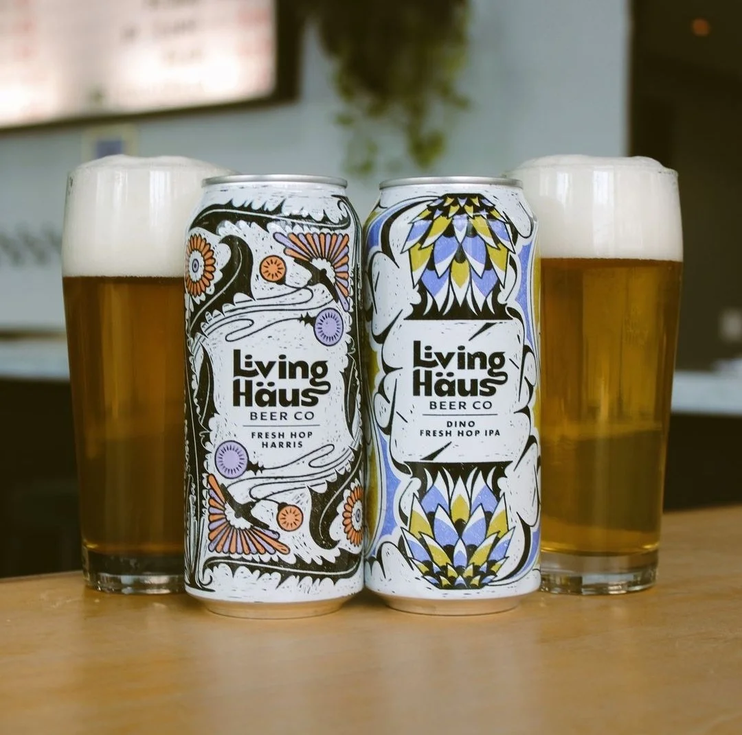 Two cans of Living Haus Beer Co. craft beer, labeled Fresh Hop Harris and Dino Fresh Hop IPA, placed between two pint glasses of beer with foamy heads on a wooden table.